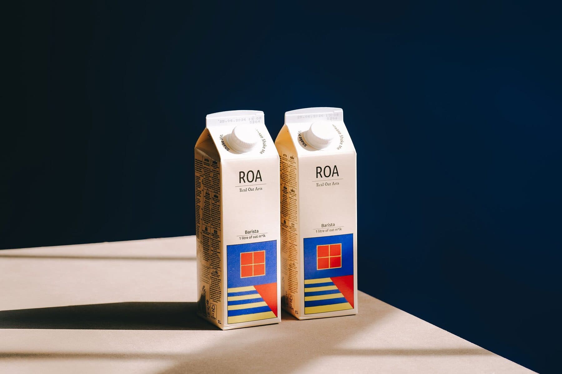 Two cartons of ROA barista oat drink on a pale surface against a deep navy wall